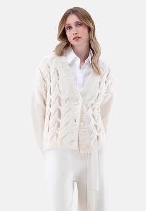 Strickjacke in Creme mit einem strukturierten Rautenmuster, V-Ausschnitt und Knopfverschluss. Kombiniert mit einem weißen Hemd und passenden Hosen.