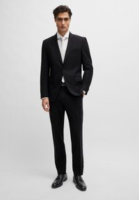 Svart dress med enkelt knappelukking, slim fit, og to frontlommer. Hvit skjorte og slips, komplementert av polerte svarte sko.