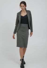 Blazer à carreaux noir et blanc et jupe crayon, associé à un haut avec bordure en dentelle noire et des bottines noires, sur un fond clair.