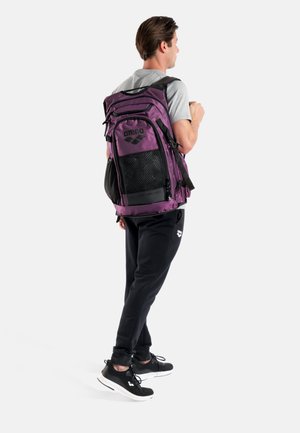 Paarse en zwarte rugzak met mesh-accenten en logo, voorzien van meerdere compartimenten en verstelbare riemen. Draagt door een persoon die van je weg staat.