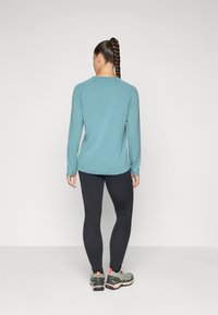 Lange mouwen, teal sporttop met een textuurpatroon, gecombineerd met zwarte leggings en grijze sneakers met oranje accenten.