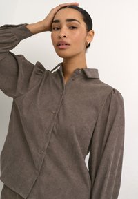 Brown button-up shirt with a soft, textured fabric, a collar, and puffed sleeves. Features a diagonal cut and front button closure.