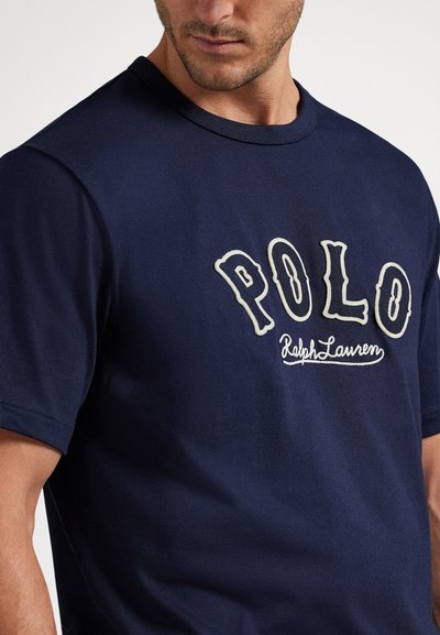 Moški, ki nosi temno modro srajco s kratkimi rokavi in velikim belim napisom "POLO Ralph Lauren" na prsih, prikazan od vratu do pasu.