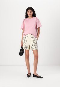 Haut rose clair à manches courtes avec des épaules volantes, associé à un short crème avec des motifs abstraits noirs et des ballerines noires. Petit sac à main noir.
