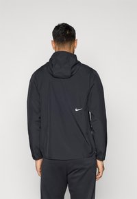 Μαύρο αδιάβροχο μπουφάν Nike με κουκούλα, ομαλή υφή, ελαστικές μανσέτες και λευκό λογότυπο στην πλάτη. Μινιμαλιστικός σχεδιασμός.