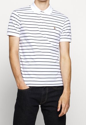 Mężczyzna w białej koszulce polo w czarne poziome paski z małym haftowanym logo, w połączeniu z ciemnoniebieskimi dżinsami, stoi z jedną ręką w kieszeni.