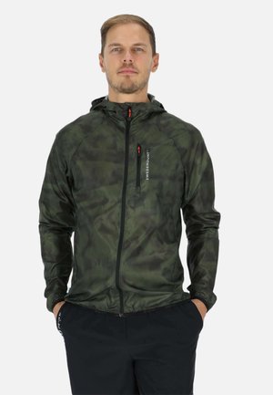 Man wearing a green camo hooded zip-up jacket with hands in pockets, standing against a plain white background.