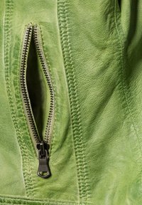 Grüne Lederjacke mit einer Reißverschlusstasche, sichtbaren Nähten und einer strukturierten Oberfläche, die das Material und die Verarbeitung des Kleidungsstücks betont.