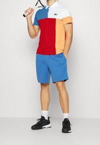 Męska koszulka polo z krótkim rękawem w czerwonym, niebieskim, białym i pomarańczowym wzorze color-block. Połączona z niebieskimi szortami i czarnymi butami sportowymi.