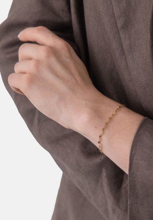 Handgelenk mit zartem goldenen Kettenarmband, getragen über einem braunen Langarmshirt, Hand ruht sanft auf dem gegenüberliegenden Arm.