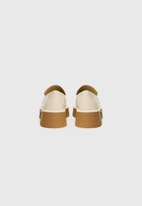 Gai bēša krāsa slip-on kurpes ar brūniem akcentiem; gluda materiāla, noapaļota forma un masīvi zoli ar horizontālām rievojumiem.