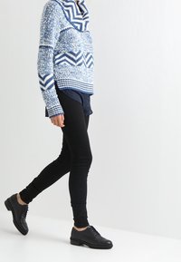 Suéter de punto con patrón de sargado en azul y blanco, con cuello de barco, llevado sobre una camisa oscura, combinado con jeans ajustados negros y zapatos negros con cordones.