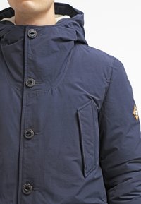 Granatowa, ocieplana kurtka z wysokim kołnierzem, z zapięciem na guziki i kieszenią boczną. Posiada fakturowaną tkaninę oraz naszywkę na ramieniu.