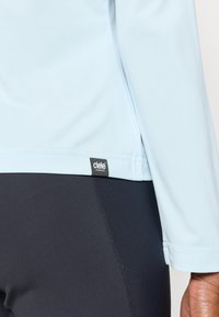 Hele sinine pika varrukaga särk, valmistatud sileda kangaga, koos tumedate legg-insidega. Särgil on väike must logo sildi servas.