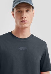 T-shirt en coton gris foncé à manches courtes ; présente un texte subtil au centre indiquant "JE ME MOQUE DE TOUT, JE ME DÉTENDS."