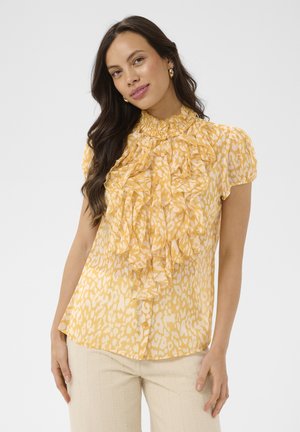 Kobieta z długimi ciemnymi włosami, nosząca złote kolczyki koła oraz żółtą bluzkę w wzory z falbanami z przodu i bufiastymi rękawami, stoi na białym tle.