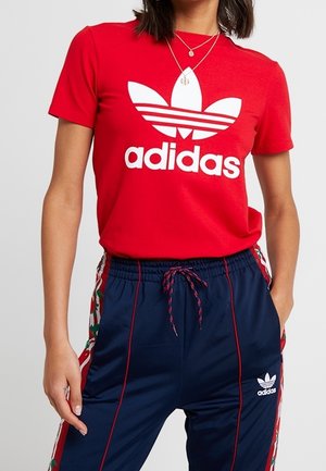 Nainen, jolla on yllään punainen Adidas-t-paita valkoisella logolla sekä tummansiniset Adidas-verryttelyhousut, joissa on kukkakuviolliset sivuraidat ja punaiset koristeet.