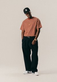 Άνδρας φορώντας μαύρο καπέλο NY, κόκκινη ριγέ μπλούζα, μαύρα παντελόνια, λευκά παπούτσια Nike, χρυσό ρολόι και κολιέ, στέκεται μπροστά σε απλό φόντο.