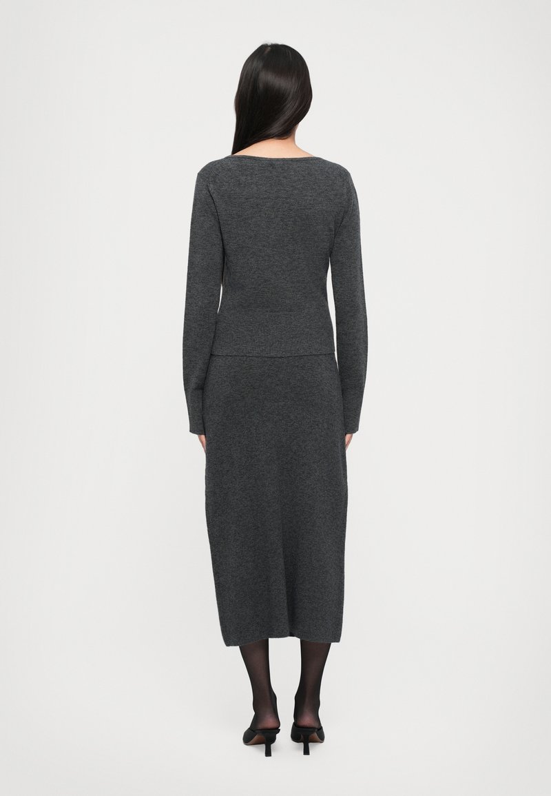 Femme avec de longs cheveux noirs, portant une robe tricotée ajustée gris foncé, des collants noirs et des chaussures à talons noires, se tenant dos à un fond blanc.