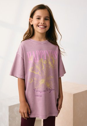Lichtroze oversized t-shirt met bloemmotieven in geel en roze. Heeft korte mouwen en een ronde hals. Draagt donkere donkerrode broek.