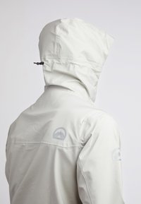 Giacca impermeabile grigia chiara con cappuccio, con un logo sul retro e sulle maniche. Tessuto liscio con un design minimalista.
