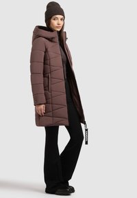 Bruine, gewatteerde pufferjas met een capuchon, met een ritssluiting aan de voorkant, zijvakken en een merketiket. Gepaard met zwarte broek en laarzen.