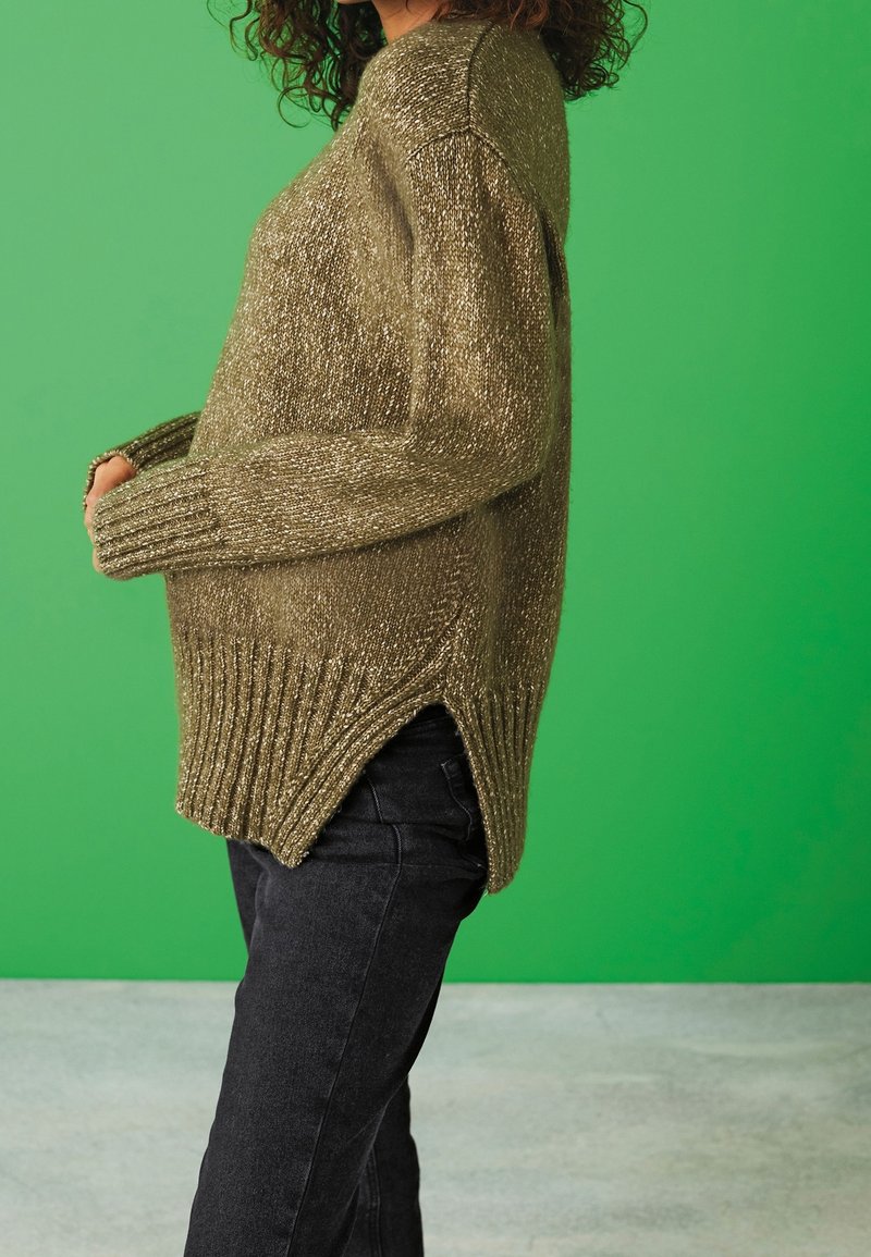 Personne portant un pull en maille vert olive avec poignets et ourlet côtelés, associé à un jean noir, debout devant un fond vert.