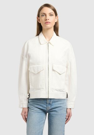 Fiatal nő fehér cipzáras dzsekiben, nagy első zsebekkel és világoskék farmerral, egyszerű szürke háttér előtt állva.