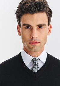 Czarny sweter z dekoltem w serek na białej koszuli, z wzorzystym krawatem w odcieniach niebieskiego, szarego i białego. Gładka faktura, dopasowany krój.