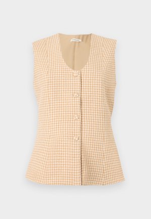 Femme portant un gilet beige à carreaux sur une blouse blanche à larges poignets, pantalon large foncé et chaussures marron, debout devant un fond uni.