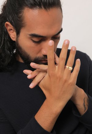 Gold-toned ring with textured band, worn on fingers, hands clasped in front. Dark ribbed sweater, soft skin texture, neutral background.