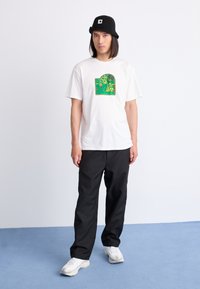 White short-sleeve t-shirt featuring a green graphic design, paired with black pants and white sneakers. Model wears a black bucket hat.
