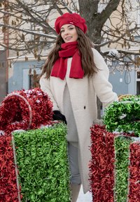 Donna in piedi all'aperto tra due scatole regalo decorate con tinsel rosso e verde, indossando un cappotto bianco, un berretto rosso e una sciarpa abbinata.