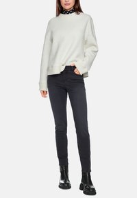 Weißer, strukturiert gestrickter Pullover mit überschnittenen Schultern, kombiniert mit schwarzen, schmal geschnittenen Jeans und schwarzen Stiefeletten. Der Pullover hat einen gerippten Saum.