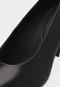 Μαύρο δερμάτινο παπούτσι με ψηλό τακούνι, στρογγυλεμένη μύτη και λείο φινίρισμα, εμφανιζόμενο σε κοντινή κεκλιμένη όψη.