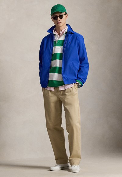 Μπλε ελαφρύ μπουφάν, πράσινη και λευκή ριγέ πόλο, ανοιχτό ροζ πουκάμισο από κάτω, μπεζ παντελόνι, λευκά αθλητικά παπούτσια, πράσινο καπέλο και γυαλιά ηλίου.