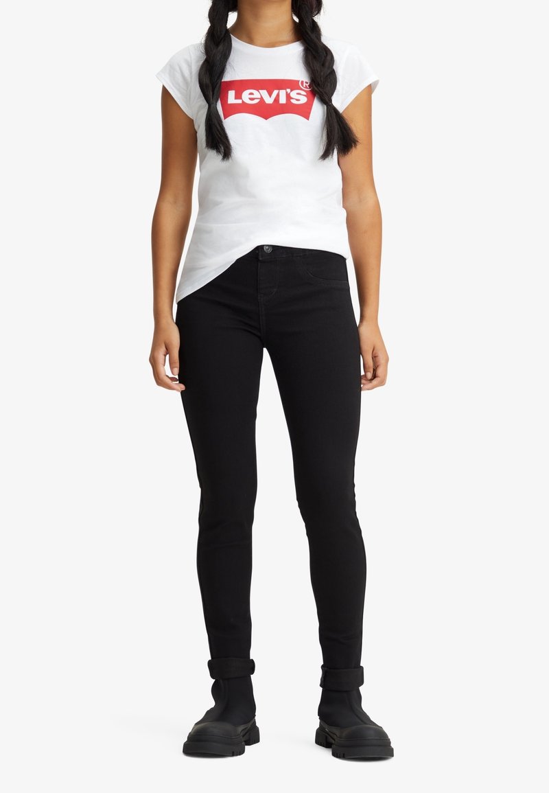 Personne aux longs cheveux tressés portant un t-shirt blanc Levi’s, un jean noir skinny et des bottes noires, debout devant un fond uni.