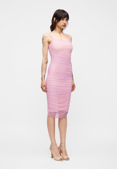 Γυναίκα με σκουρόχρωμα μαλλιά που φοράει μίντι ροζ φόρεμα χωρίς μανίκια με ρυτιδωτή υφή και μπεζ ψηλοτάκουνα σανδάλια, στέκεται μπροστά σε ένα απλό λευκό φόντο.