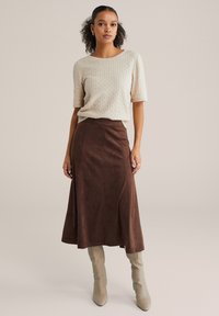 Béžový texturovaný sveter s krátkymi rukávmi v kombinácii s dlhou hnedou semišovou sukňou a svetlosivými čižmami sahajúcimi po kolená. Hladký, elegantný vzhľad.