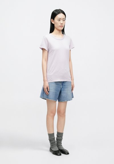 Mulher com cabelo preto liso usando uma t-shirt lilás clara, calções de ganga, meias cinzentas e sapatos planos pretos, em pé sobre um fundo branco.