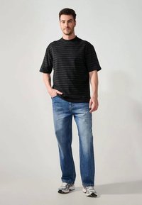 Mężczyzna stoi w czarnej, w paski, krótkiej koszulce, granatowych dżinsach i białych trampkach na jasnym, jednolitym tle.