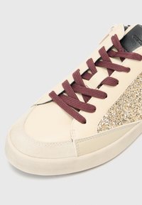 Sneaker beige in pelle con lacci bordeaux, punta in suede e pannello laterale glitterato in oro. Presenta un accento nero sul tallone.