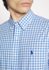 Blå och vit rutmönstrad skjorta med knäppkrage. Har pärlknappar och en liten broderad logotyp på bröstet. Avslappnad design.