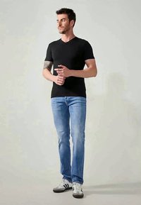 Black V-neck t-shirt, short sleeves, paired with light blue jeans and white sneakers with black accents. Model stands with hands positioned.