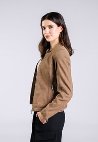 Giacca in suede marrone con design strutturato, tasche con zip e superficie liscia. Presenta un colletto rialzato e una siluetta aderente.