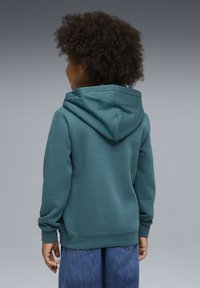 Hoodie bleu sarcelle à texture douce, doté d'une grande capuche et de poignets, associé à un jean bleu ample, vu de dos.