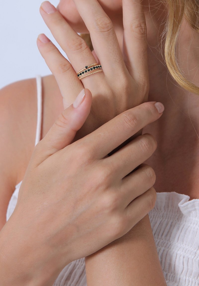 Main avec deux bagues en or, dont une avec des pierres noires, tenue délicatement par l’autre main sur un haut blanc sans manches.