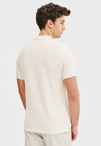 Beżowa koszulka polo z krótkim rękawem, z płaskim kołnierzykiem, gładką fakturą i subtelnymi detalami w szwie, zaprezentowana od tyłu na neutralnym tle.