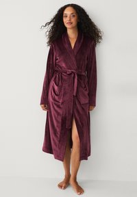 Vrouw met krullend haar, gekleed in een lange, pluche bordeauxrode badjas die om de taille is geknoopt, staande op blote voeten tegen een eenvoudige lichte muur.