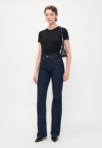 Woman standing, wearing black fitted t-shirt, dark blue high-waisted jeans, black pointed shoes, and carrying small black shoulder bag.